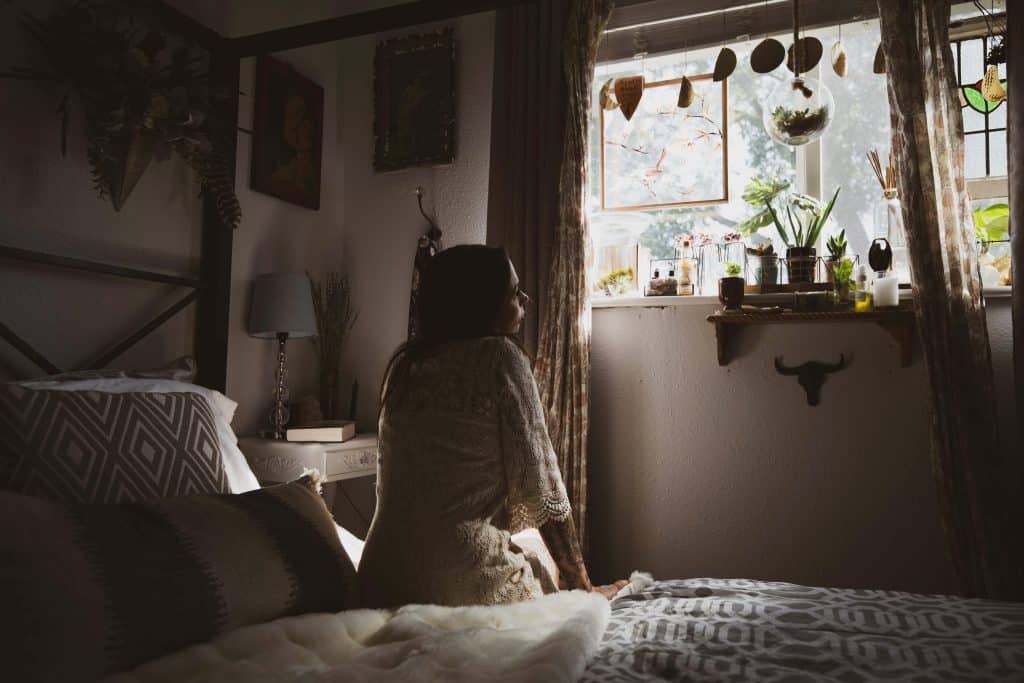 Woman sitting on her bed looking toward a sunlit window in a cozy bedroom